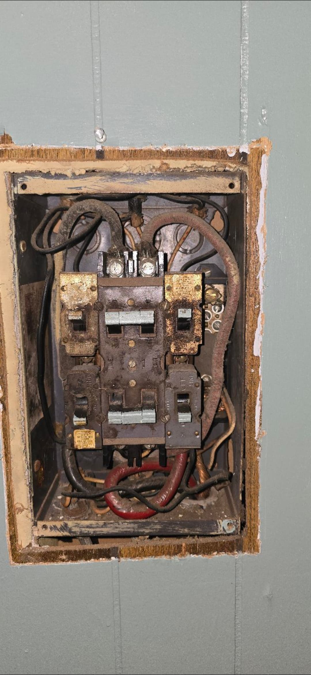 Old, dusty electrical circuit breaker panel with exposed wiring recessed into a light blue wall.
