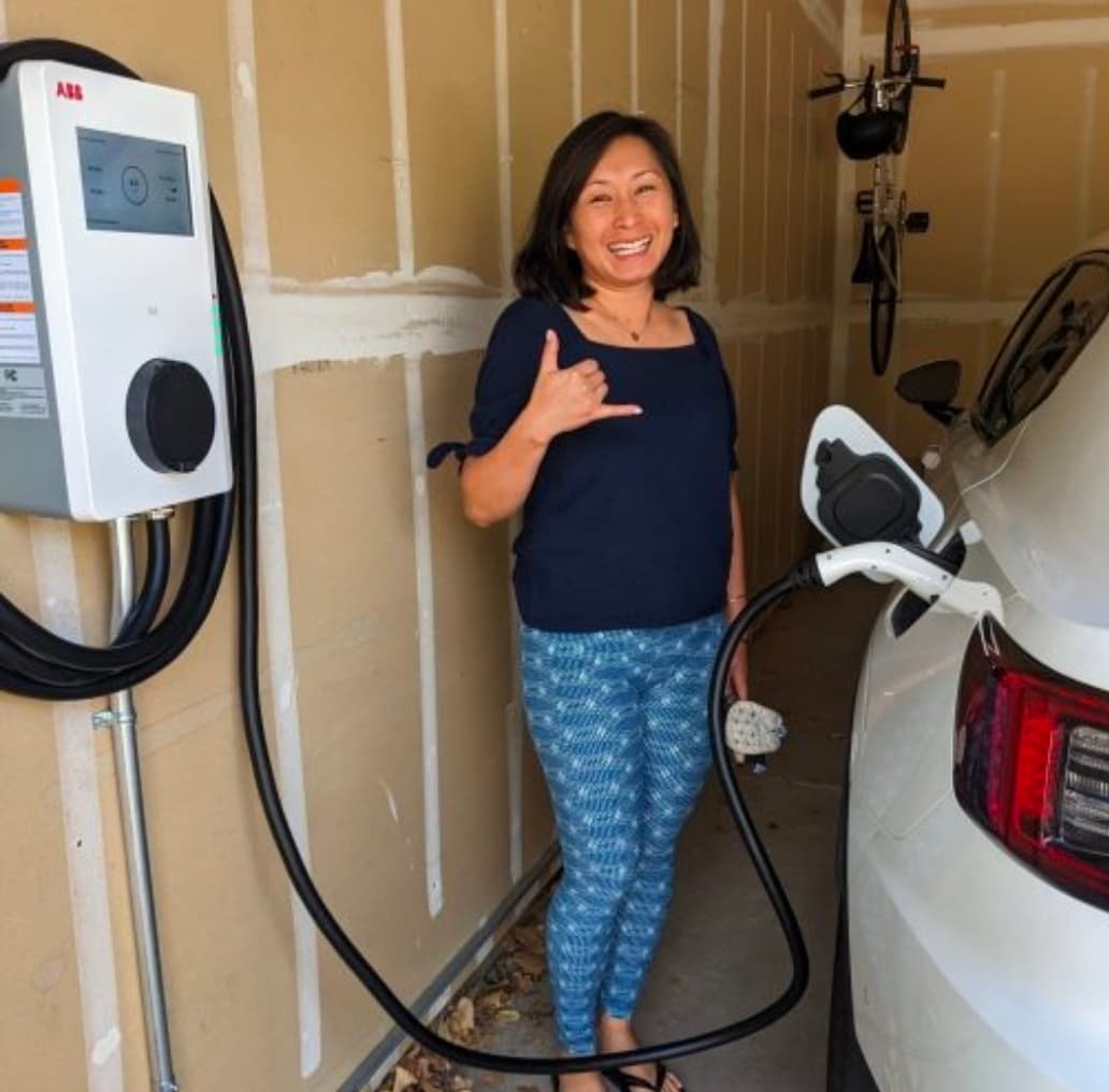 Smiling woman makes a shaka sign while charging her white electric car in a garage.