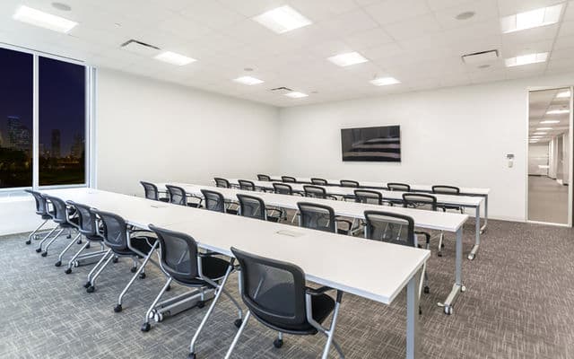 Modern training room with rows of white tables, black mesh chairs, and a wall-mounted monitor.