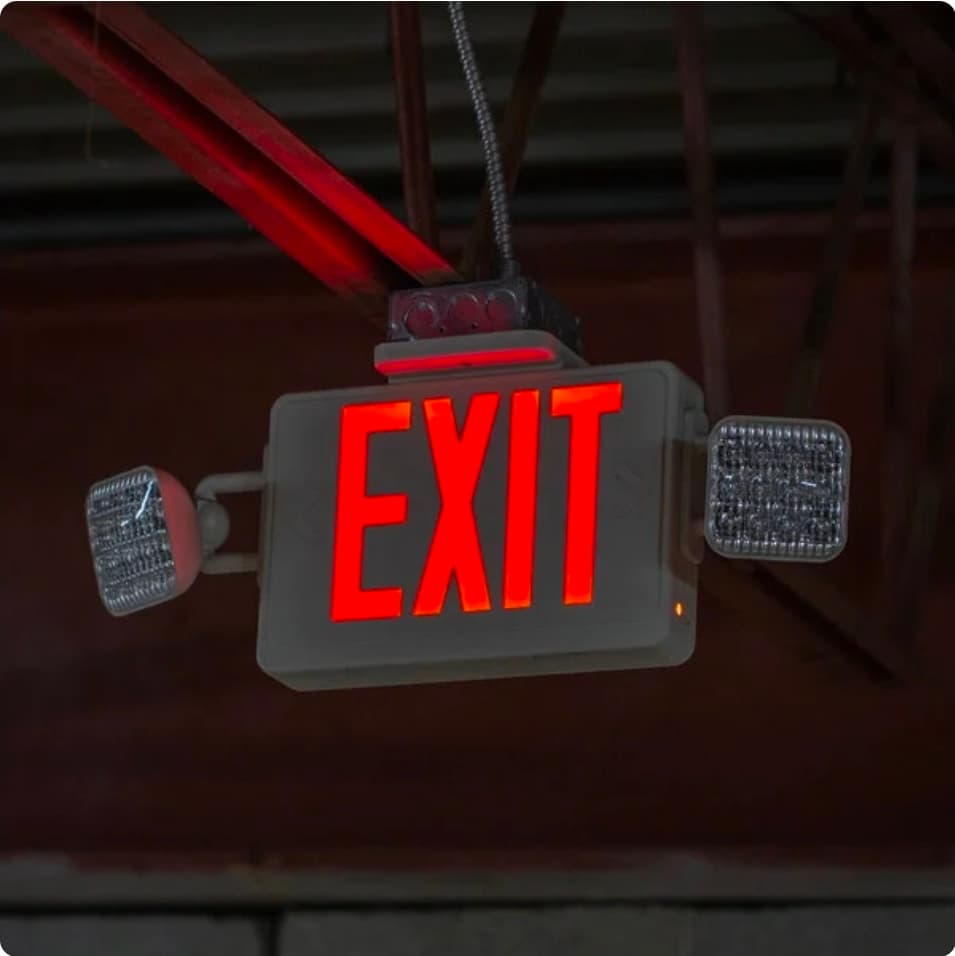 Illuminated red EXIT sign with emergency lights hanging from a red industrial ceiling beam.